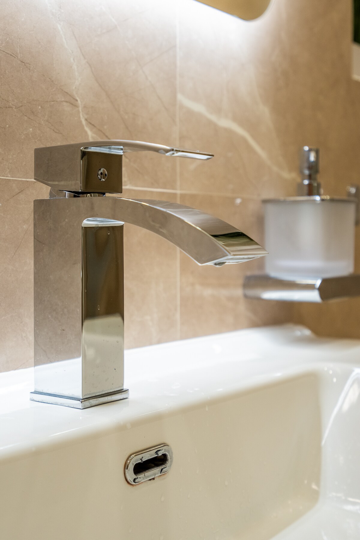 A modern bathroom sink features a sleek chrome faucet and a minimalist soap dispenser mounted on the wall. The sink is surrounded by smooth, beige marble tiles, providing an elegant backdrop to the contemporary fixtures.