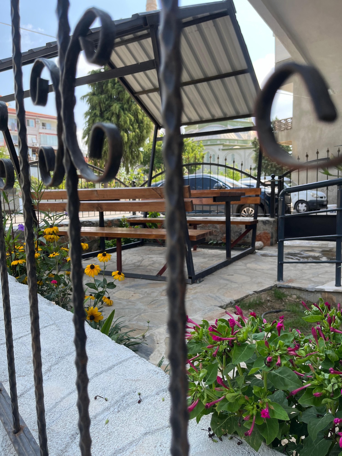 A garden area is depicted, featuring a shaded wooden gazebo with tables and benches, surrounded by vibrant flowers. A stone pathway leads to the seating area, and a decorative iron gate provides access. The view highlights a blend of nature and outdoor relaxation.