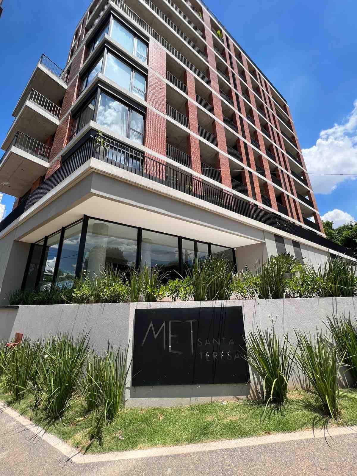 The exterior of a contemporary building is showcased, featuring a mix of brick and glass. Large windows dominate the ground level, providing natural light to the interior. Lush greenery encircles the entrance, with a prominent sign displaying the name of the building.