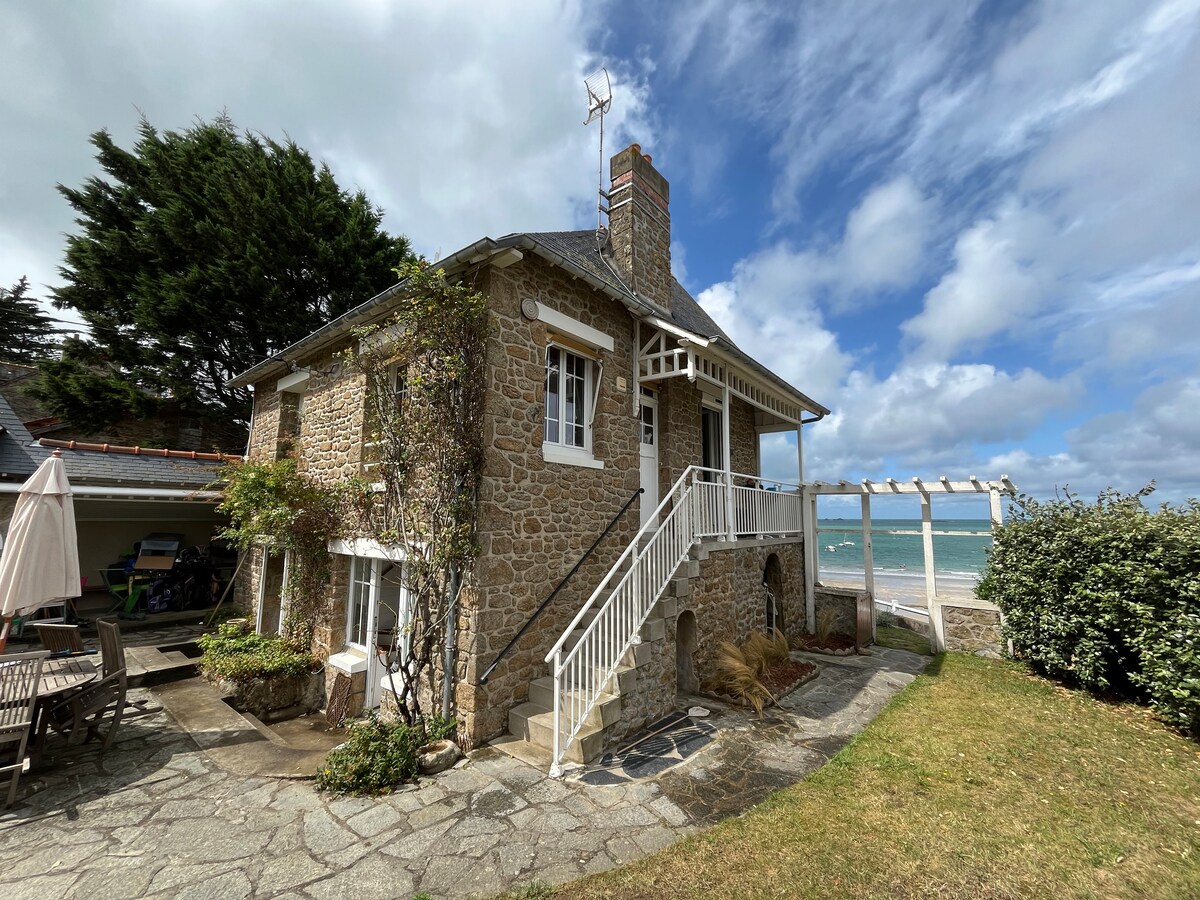 The exterior of a stone house is displayed, featuring traditional architecture with a staircase leading to a covered terrace. Lush greenery surrounds the property, and the nearby beach and sea are visible in the background under a partly cloudy sky.