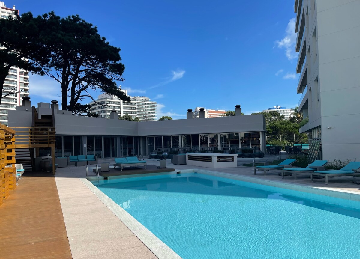 An outdoor swimming pool is surrounded by lounge chairs and seating areas. The pool reflects the clear blue sky above, while nearby buildings are visible in the background. A wooden deck leads up to the pool area, providing a welcoming space for relaxation.