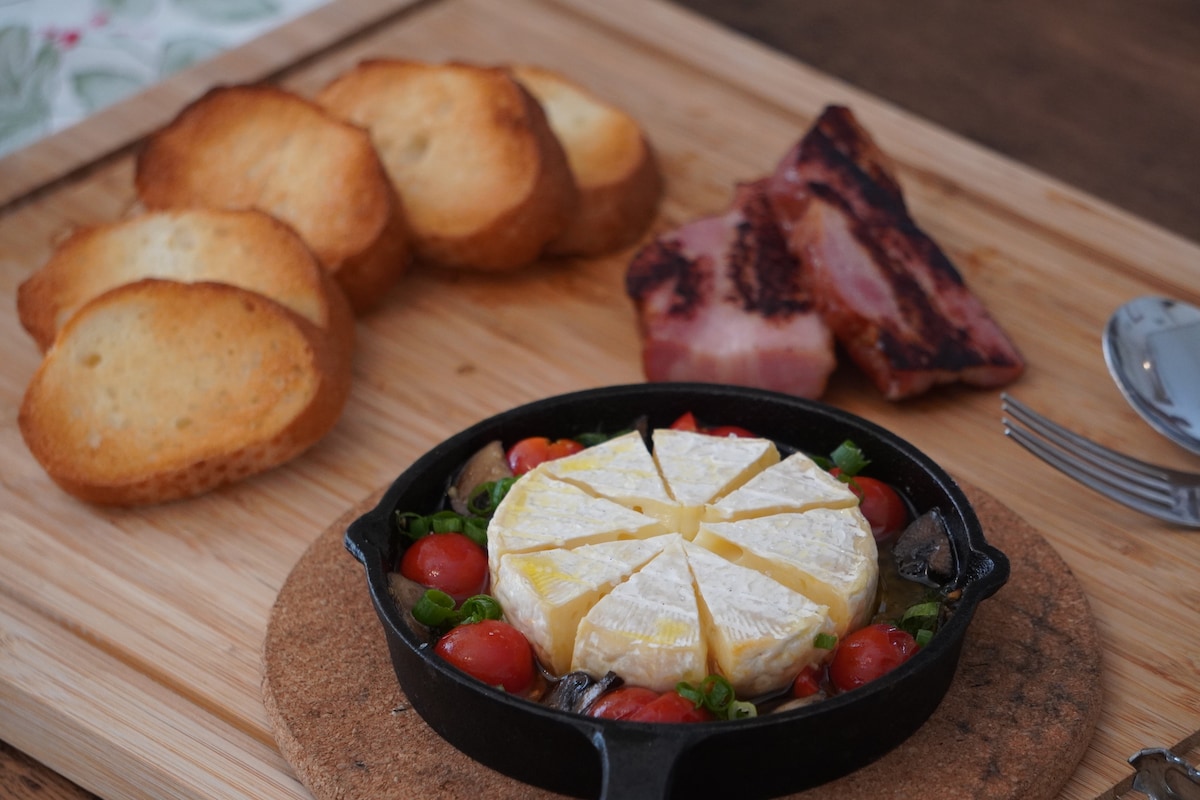 A cast-iron skillet holds a baked cheese surrounded by cherry tomatoes and green onions. Next to the skillet, toasted slices of bread and pieces of cooked bacon are arranged on a wooden cutting board. A fork and knife are positioned nearby.