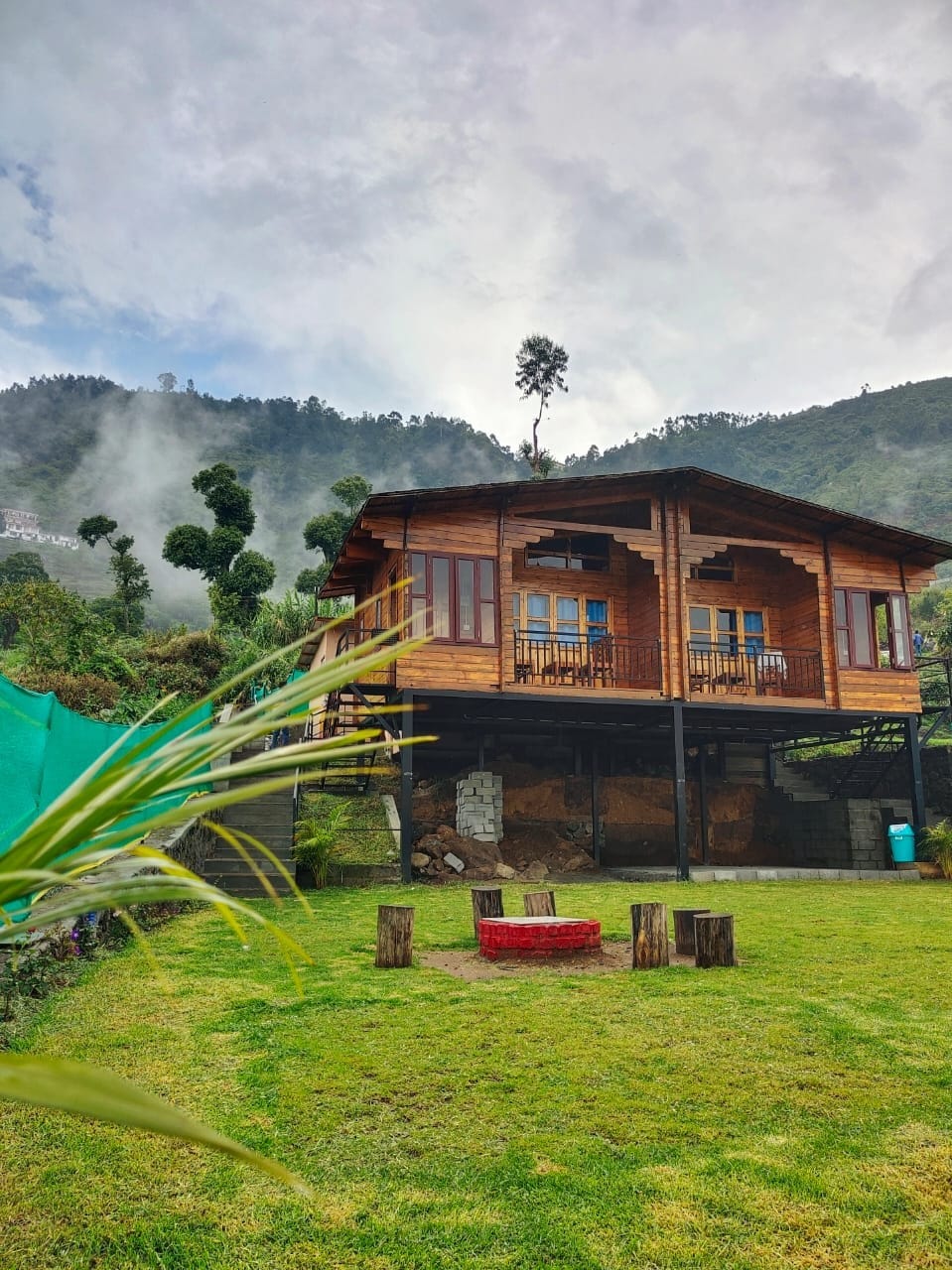 A wooden cabin structure is situated on elevated ground, surrounded by lush greenery and mist-covered hills. Two balconies are visible, adorned with chairs, offering scenic views. Below, a circular stone fire pit is surrounded by natural seating made of logs, set in a grassy area.