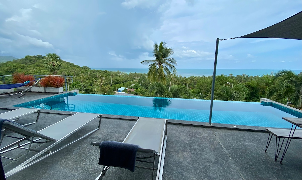 An infinity pool extends towards the horizon, surrounded by a spacious sun terrace. Lounge chairs are positioned for relaxation, offering views of lush greenery and the ocean beyond. A shaded area provides comfort from the sun, enhancing the tranquil atmosphere.