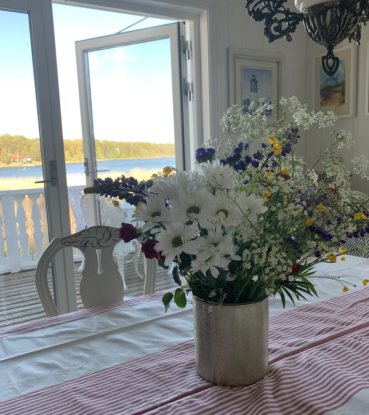 A dining table is set with a striped tablecloth, featuring a vase of fresh wildflowers. Large doors open to a scenic view of the water and nature beyond, providing a light and inviting atmosphere.