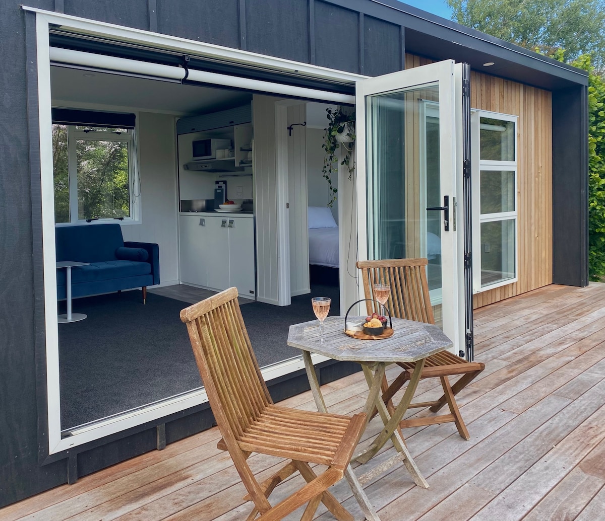 An inviting outdoor seating area features a small wooden table with two chairs, positioned on a wooden deck. Glass doors open to an interior space, showcasing a cozy sofa and a glimpse of a sleeping area. Natural light enhances the serene ambiance.
