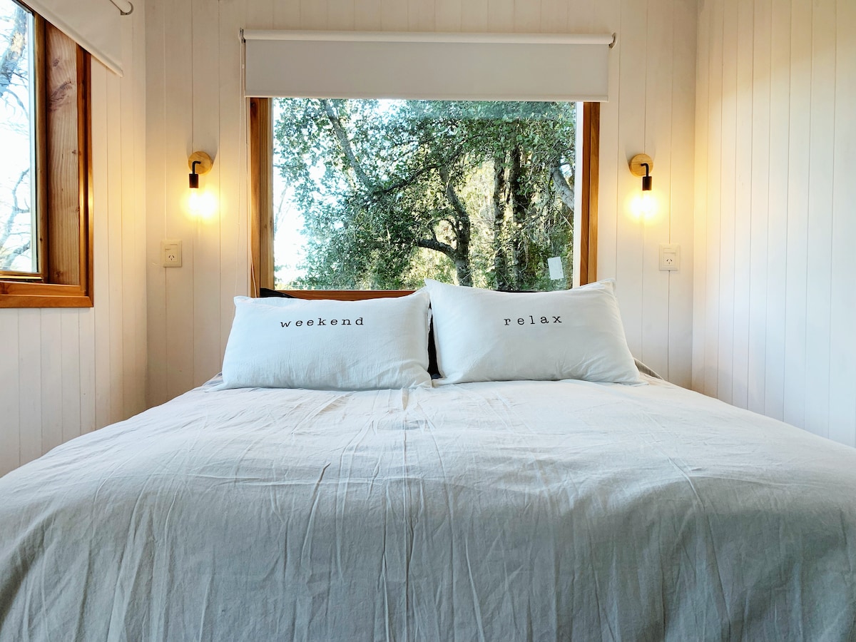 A cozy sleeping area features a spacious bed with light-colored bedding. Two decorative pillows with the words 'weekend' and 'relax' are positioned on the bed. Natural light streams through large windows overlooking lush trees, enhancing a serene atmosphere.