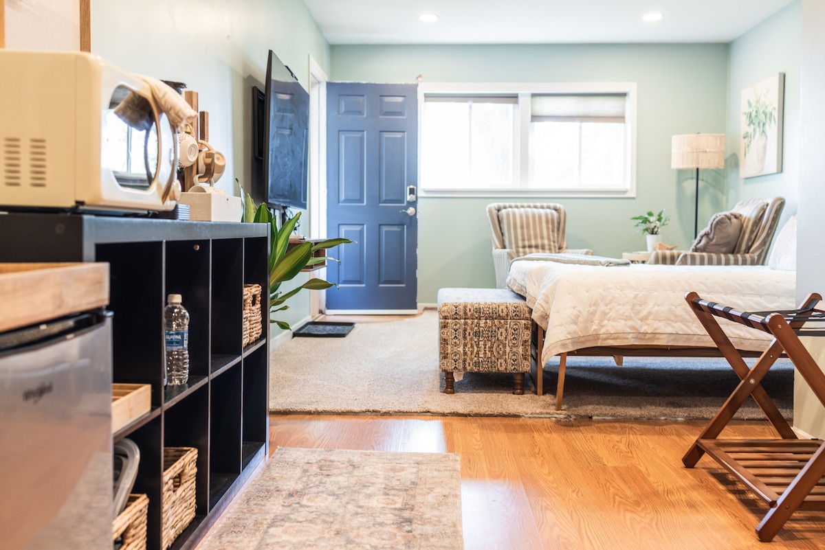 A well-appointed guest suite is visible, featuring a clean and comfortable layout. The room contains a spacious bed, a large screen TV, and a cozy sitting area with a side table. A private entrance is situated at the front, with soft natural light filtering through the blinds.