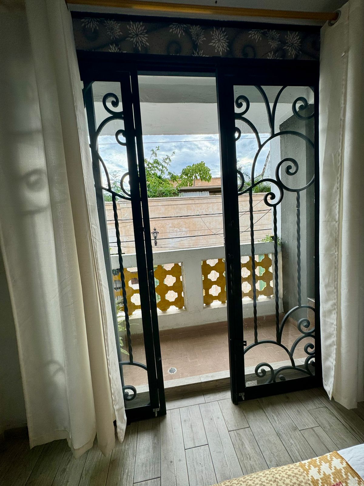 Double doors adorned with intricate ironwork open up to a private balcony. Sheer curtains frame the entrance, allowing natural light to fill the room. A view of the neighborhood can be seen beyond, framed by the architectural details.