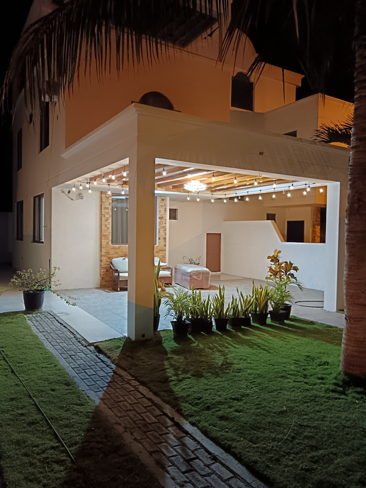 An outdoor patio is illuminated by soft lights strung overhead. Comfortable seating arrangements are visible under the structure, surrounded by lush greenery and potted plants. The space is designed to promote relaxation, with a wide pathway leading through the well-maintained lawn.