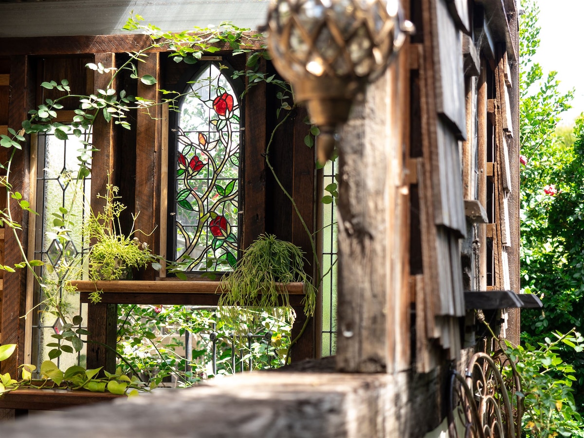 A vibrant stained glass window adorned with red roses allows natural light to filter through. Surrounding greenery spills onto wooden shelves, creating a harmonious blend of nature and artistry. The rustic wood accents add texture to the tranquil setting.