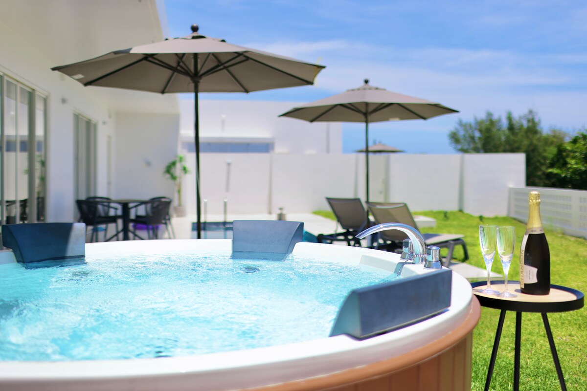 A well-maintained outdoor jacuzzi sits invitingly on a grassy area, surrounded by several lounge chairs and umbrellas. Nearby, a small table holds two champagne flutes and a bottle, suggesting a relaxing retreat under a clear blue sky.
