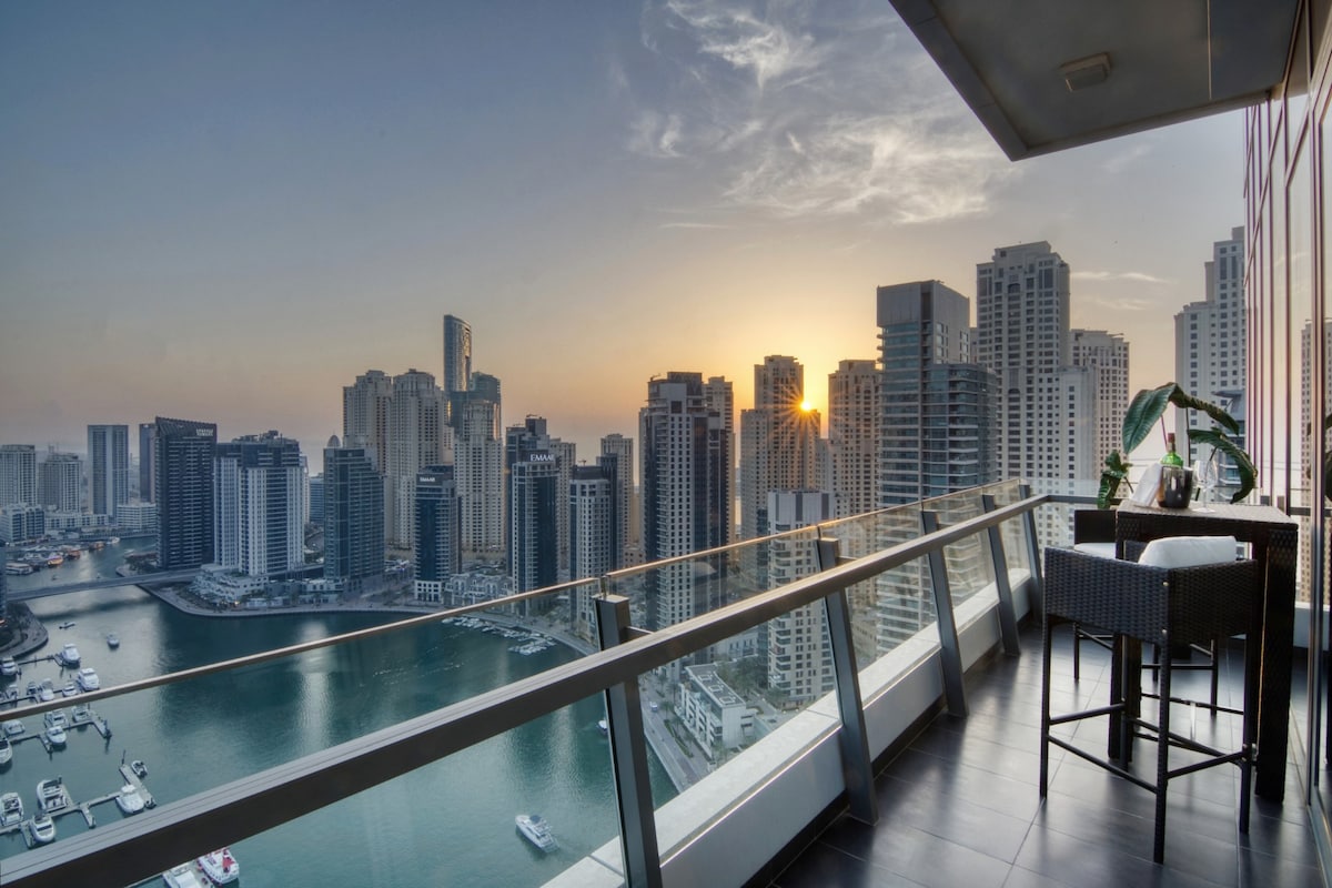 A private balcony is presented with modern outdoor seating overlooking the cityscape of Dubai Marina. The view includes a serene body of water surrounded by tall buildings, with the sun setting in the distance, casting a warm glow across the skyline.