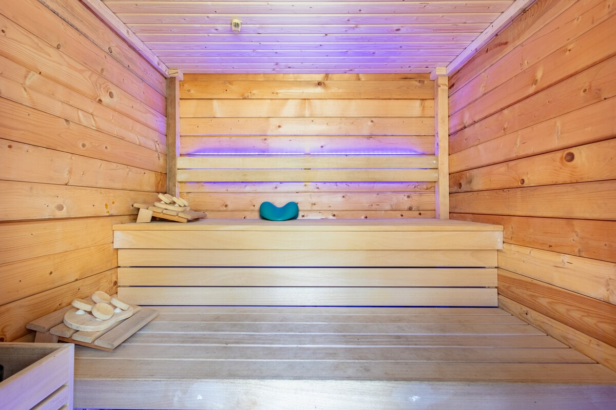 The sauna features wooden walls and benches arranged for relaxation. Ambient lighting softly illuminates the space, enhancing a calming atmosphere. Wooden accessories are placed on the side, contributing to the overall natural aesthetic.