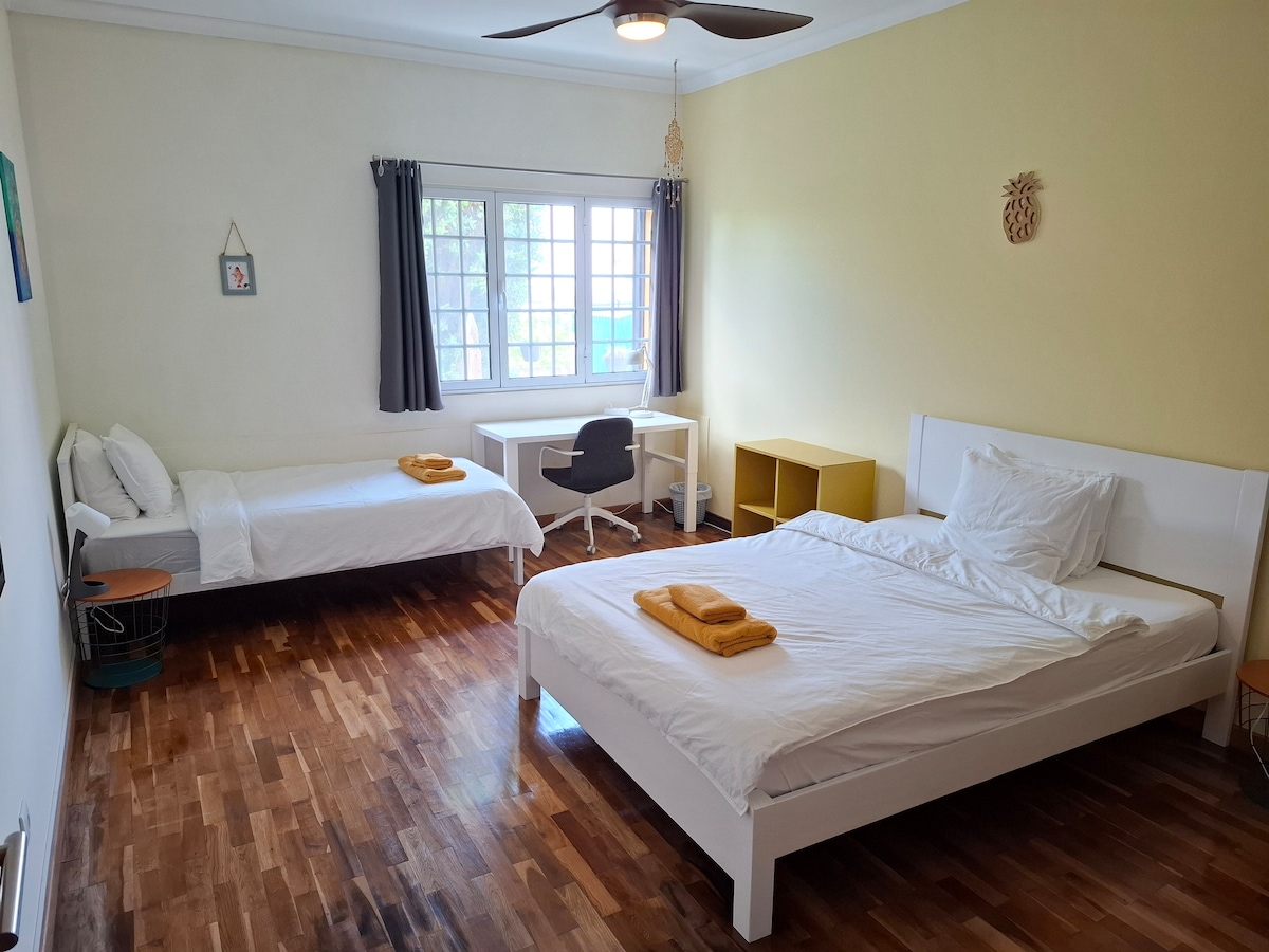 A bright room features two spacious beds, one with an additional bed beside it. A small desk and chair are positioned near a window that allows natural light, while a bedside table and a decorative wall piece add character to the space.