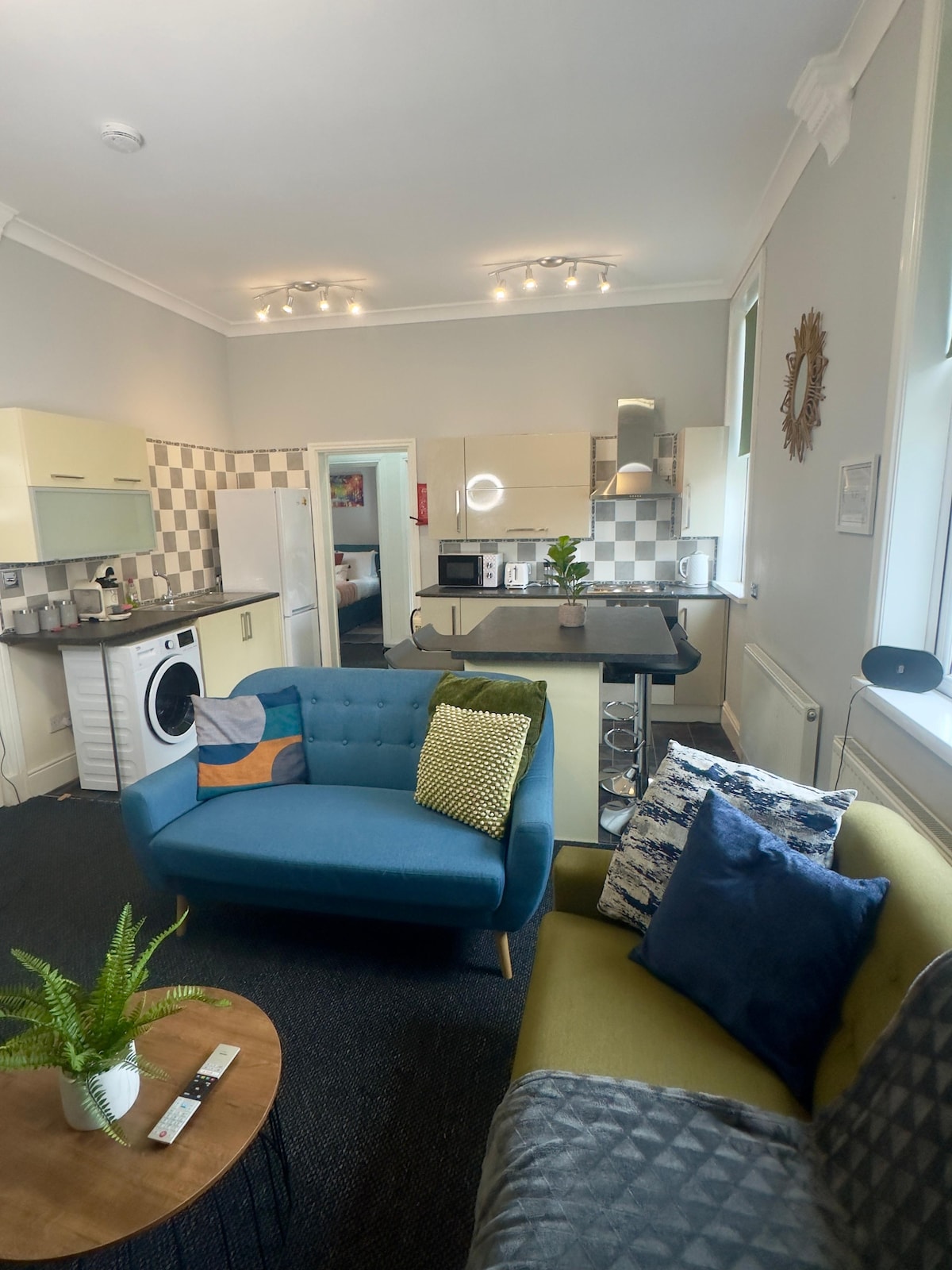 A modern open-plan living area displays a blue couch and a green chair on plush carpet. A round coffee table sits nearby, with a small potted plant atop it. The kitchen is visible in the background, featuring light cabinets and checkerboard backsplash, with a washer and dryer integrated into the design.