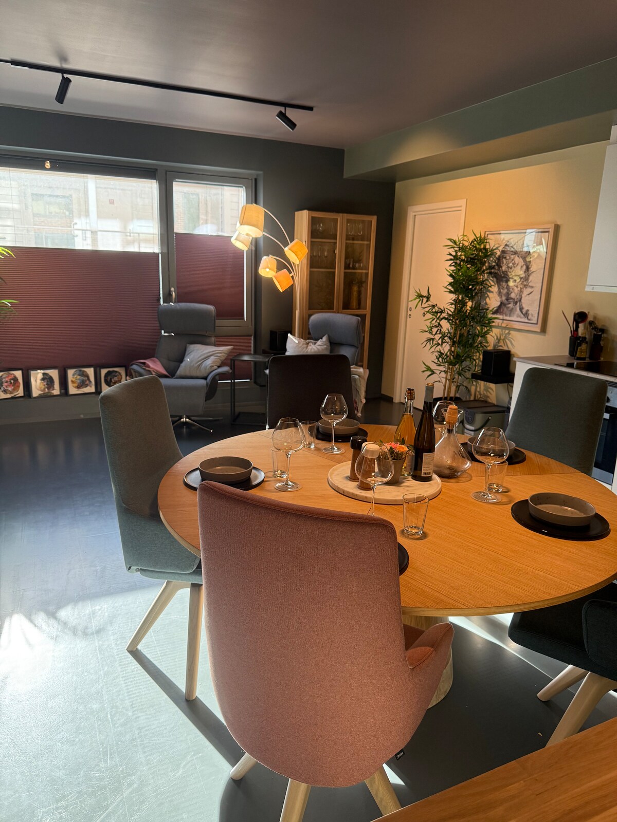 A dining area features a round wooden table surrounded by six lightly colored upholstered chairs. In the background, a cozy seating area is visible with two armchairs and a console cabinet. Natural light is filtered through large windows, enhancing the inviting environment.