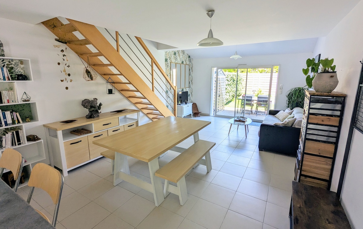 An open living area is shown, featuring a large dining table made of light wood, surrounded by chairs. A staircase with wooden steps leads to the upper level. Glass doors open to a patio with outdoor seating, allowing ample natural light into the space.