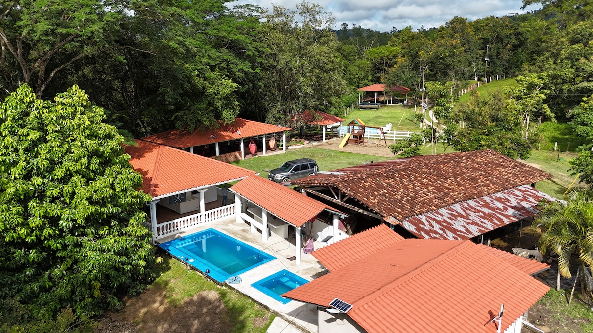 An aerial view of Casa Sinclair showcases the inviting swimming pool surrounded by lush greenery. Several red-roofed buildings are visible, including a barbecue area and outdoor seating. The property features open spaces, well-maintained lawns, and scenic landscaping, creating a tranquil outdoor environment.