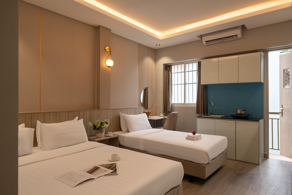 This modern studio features a spacious layout with a large double bed and a twin bed. A small kitchenette with a blue backsplash and seating area is visible. Natural light enters through a window, illuminating the neutral color palette and elegant wall fixtures.