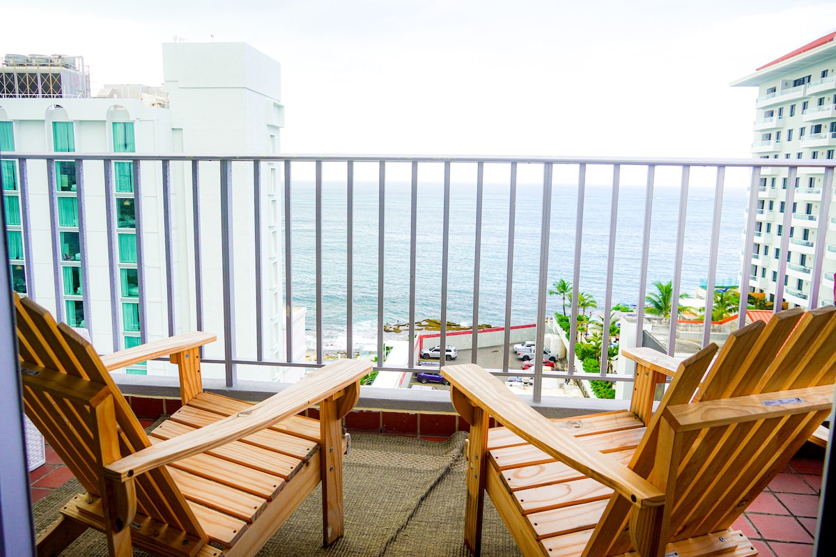 Two wooden lounge chairs are positioned on a private balcony, offering unobstructed views of the ocean. The railing frames the scene, while the distant coastline and gentle waves can be seen beyond. The ambiance is serene, enhanced by the presence of surrounding palm trees.