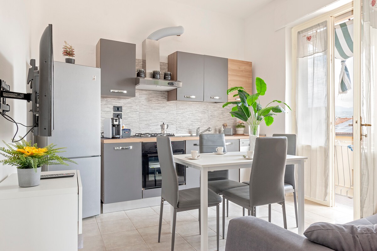 A modern kitchen is equipped with stainless steel appliances, including a refrigerator and a stove. A dining table with gray chairs provides seating for four. Natural light enters through a glass door, which leads to a small balcony, complemented by indoor plants and minimalist decor.