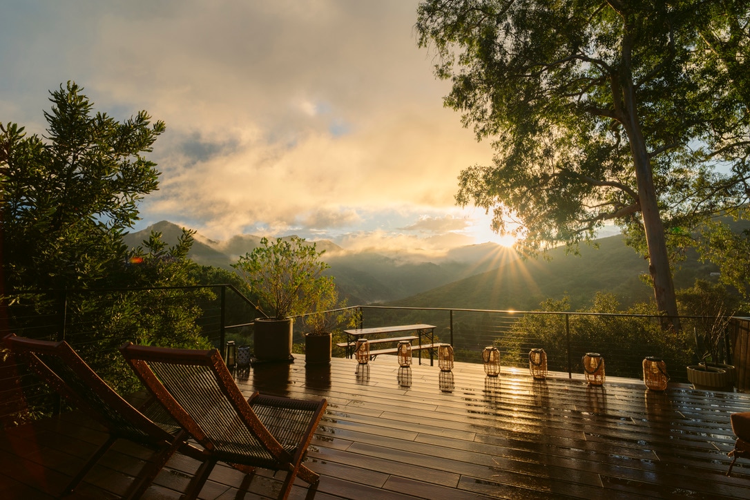 A spacious deck is showcased, featuring wooden loungers and a table overlooking lush green mountains. Sunlight streams through a cloudy sky, illuminating the scene. Decorative lanterns line the edge of the deck, creating a serene atmosphere in the early morning light.