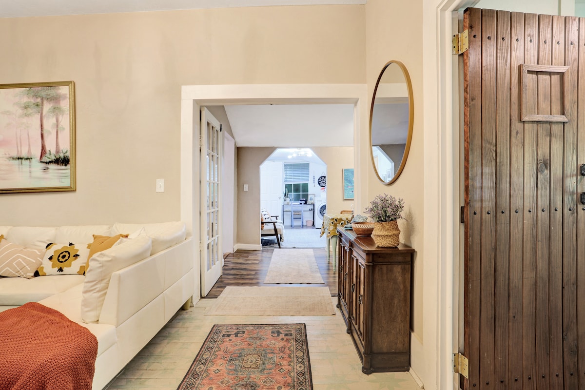 The entryway features a wooden door leading to an open space with natural light. A cozy sofa with decorative pillows is positioned nearby. A wooden console table holds a small plant, and a round mirror reflects the inviting atmosphere of the adjoining rooms.
