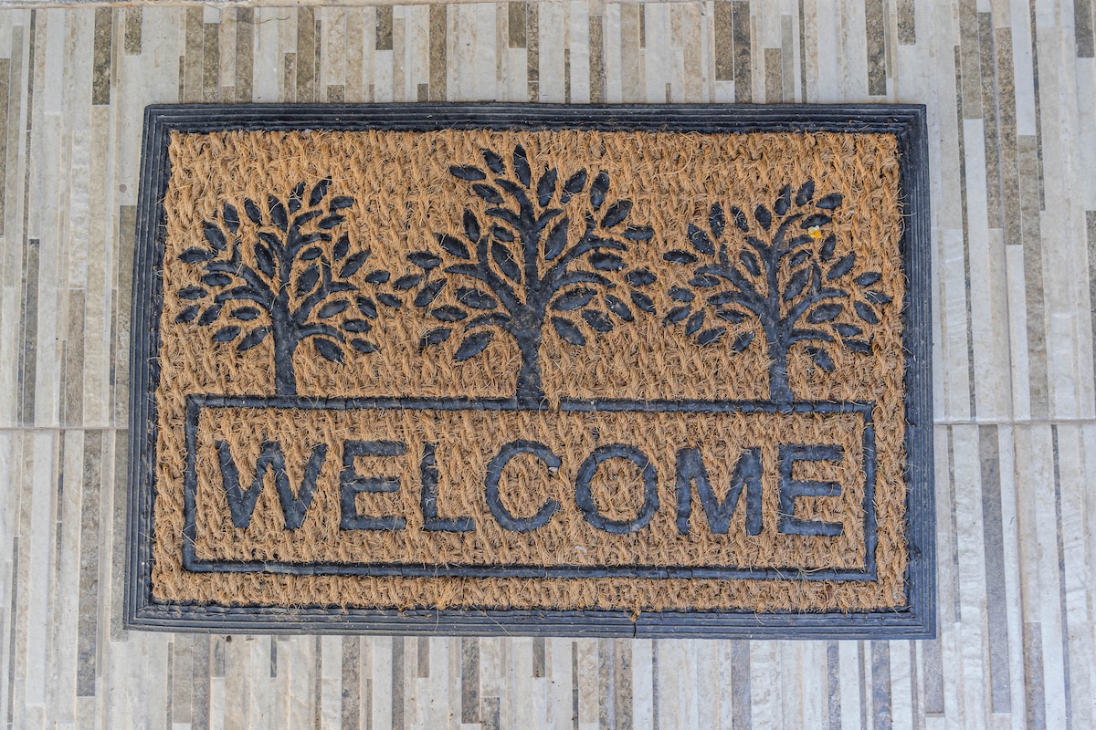 A welcome mat is displayed at the entrance, designed with a natural fiber base and featuring three tree silhouettes above the word 'WELCOME' in bold letters. The mat is positioned on a textured stone surface, creating an inviting entrance.