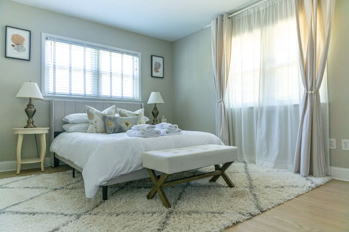 Chumba cha kulala cha malkia chenye madirisha makubwa, benchi nzuri na rangi laini zisizo na upendeleo. Inafaa kwa ajili ya kupumzika huku mwanga wa asili ukiingia na mikwaruzo ya kitambaa chini ya miguu. 