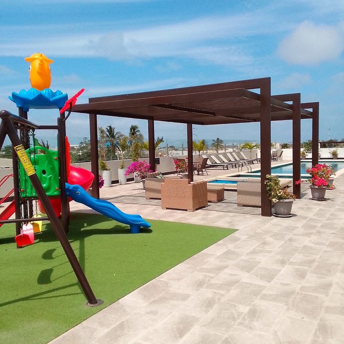 A play area features a colorful climbing structure alongside a green slide. Comfortable seating arrangements are under a shaded pergola, with lounge chairs positioned near a swimming pool. Potted flowers add vibrant touches to the outdoor space, enhancing the inviting atmosphere.