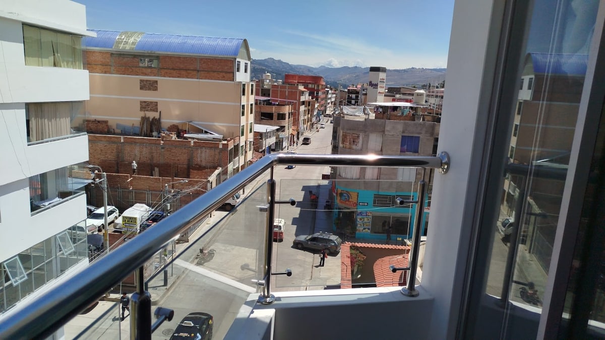 A view from a balcony overlooks a bustling street, with buildings lining both sides. Nearby, cars can be seen parked along the curb, and distant mountains create a scenic backdrop against a clear blue sky.