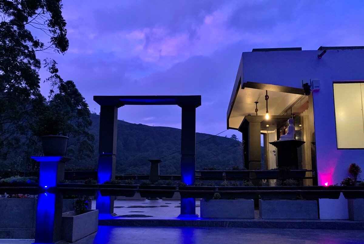 An outdoor seating area is illuminated with soft blue lighting under a twilight sky. The contemporary structure is enhanced by a large archway, framed by lush greenery, leading to a garden area. The scenic hills are visible in the background, creating a serene atmosphere.