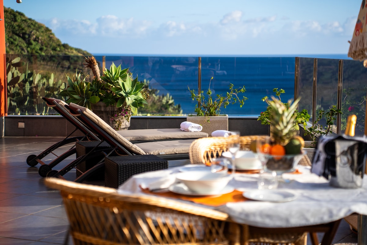 A spacious terrace overlooking the ocean features comfortable loungers and a dining area. Indoor and outdoor plants are present, complementing the scenic view. A set table with clean dishes is positioned nicely, inviting relaxation and enjoyment of the surroundings.