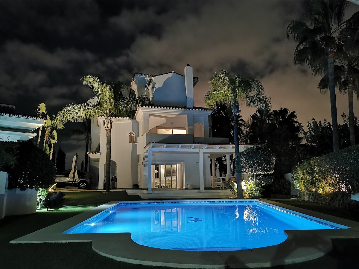 The villa is illuminated at night, showcasing its modern architecture and large glass doors. A heated pool reflects the soft blue light, surrounded by well-maintained green landscaping and palm trees. The tranquil ambiance is enhanced by the surrounding clouds in the night sky.