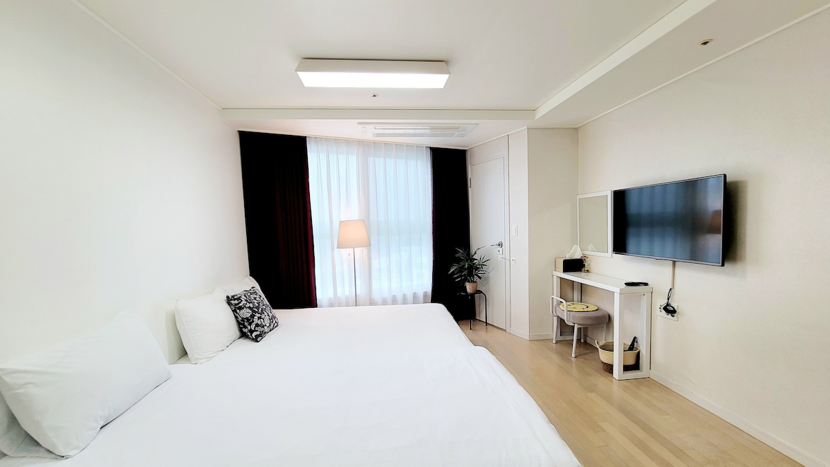 A spacious room features a large white bed on one side and a sleek desk with a television mounted on the wall across from it. Natural light enters through sheer curtains, and a floor lamp and small plant add to the minimalist decor.