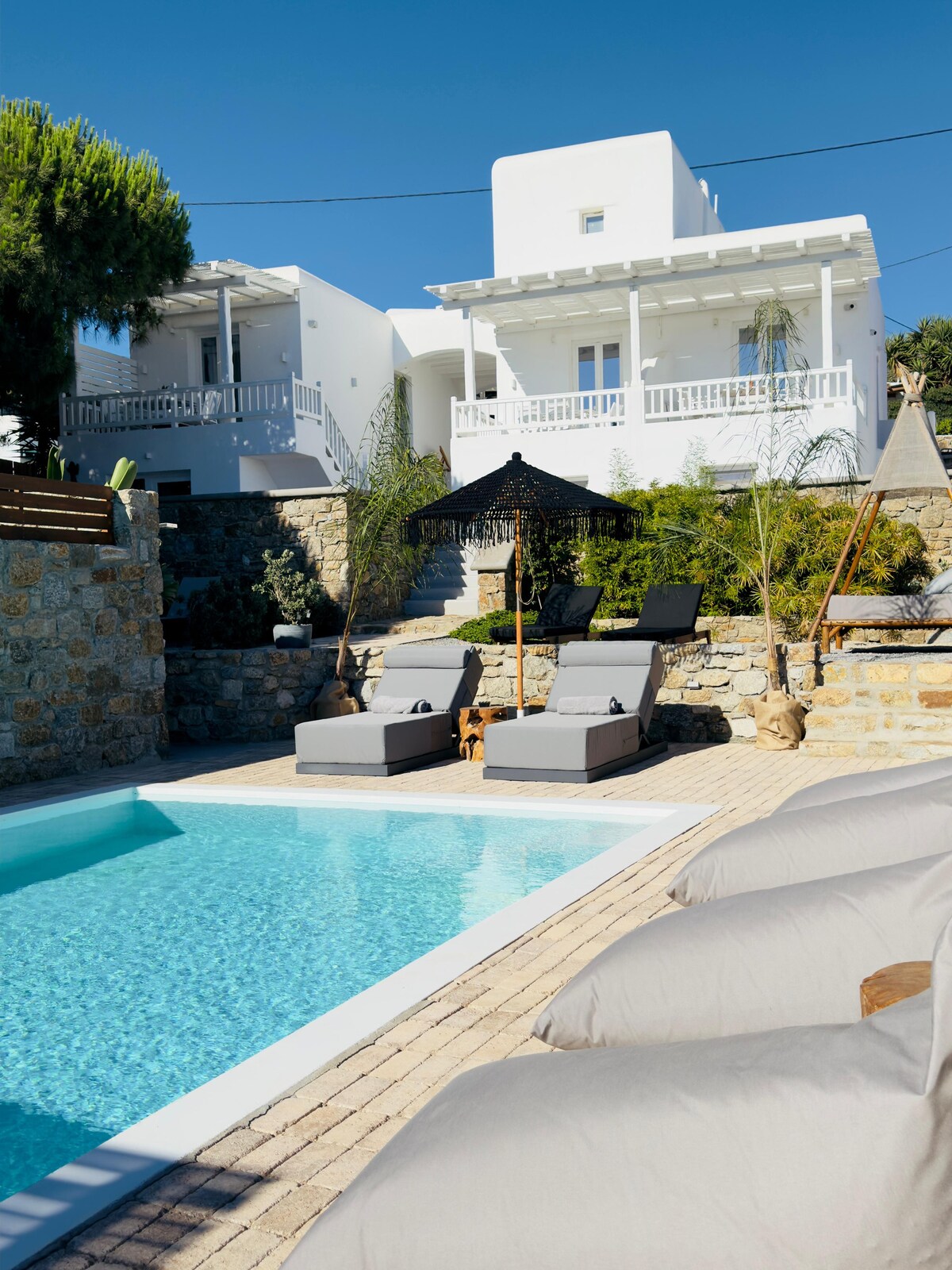 A well-maintained outdoor area features a turquoise swimming pool surrounded by comfortable lounge chairs. Sun loungers are positioned on the patio, with greenery framing the space. The white building in the background complements the serene atmosphere, creating a relaxing retreat.