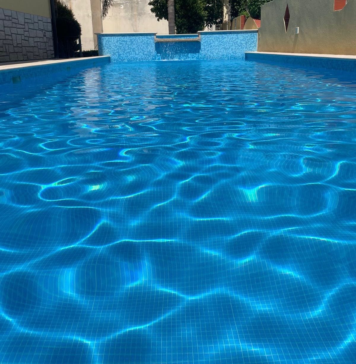 A swimming pool is filled with clear blue water, reflecting light and creating a shimmering effect on the surface. The pool's smooth edges are visible, surrounded by a tiled border, inviting relaxation and enjoyment.