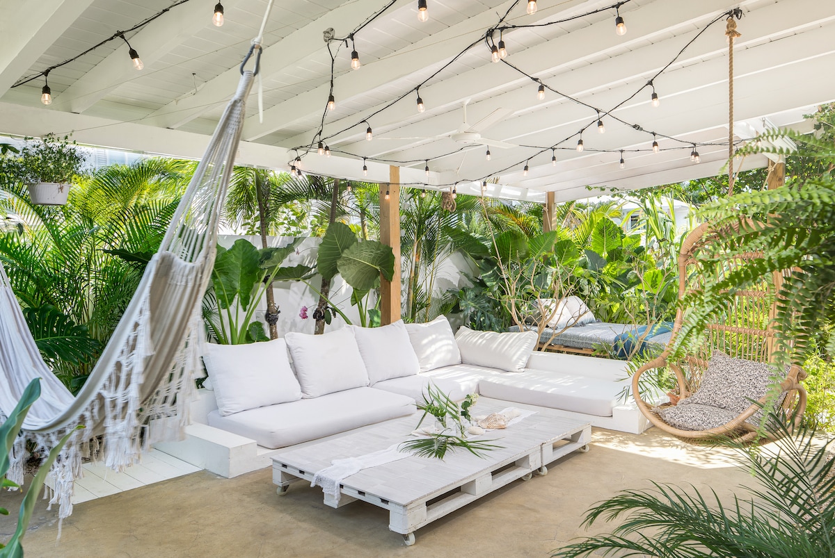 An inviting outdoor lounge area features a white sectional sofa adorned with soft cushions, surrounded by lush tropical plants. A hammock hangs nearby, and a suspended chair provides additional seating. String lights illuminate the space, creating a relaxing atmosphere near the natural pool.