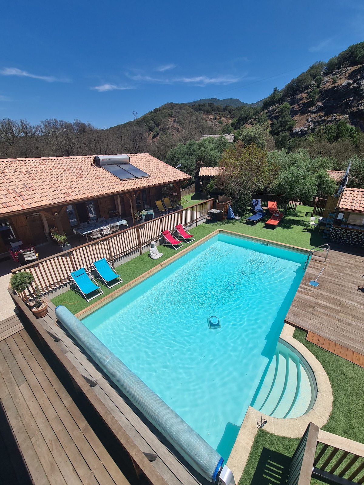 A clear blue swimming pool is surrounded by wooden decking and colorful lounge chairs. The area is enhanced by greenery and features views of the nearby mountains, creating a serene outdoor space. The sunlit building offers access to the pool area.