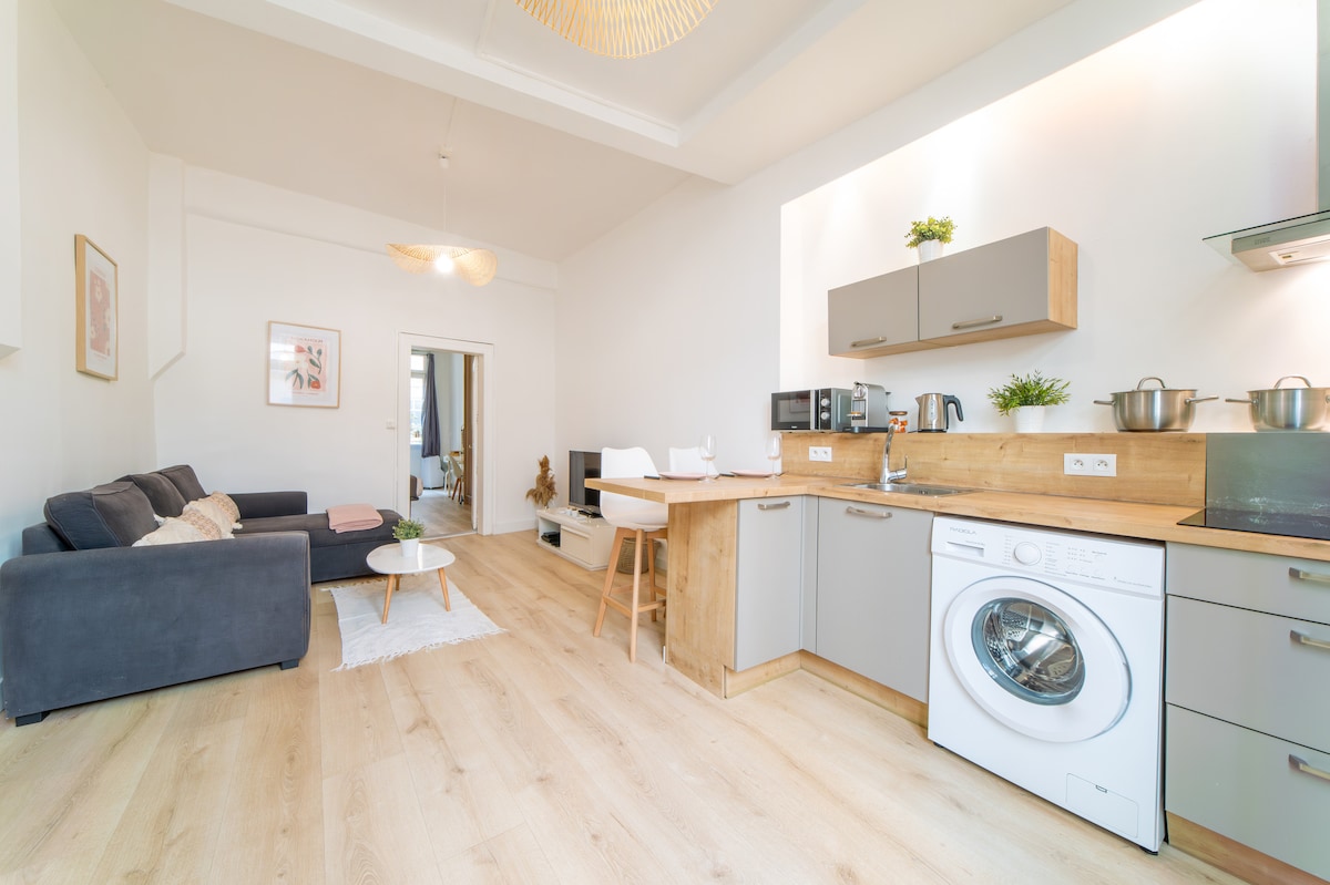 An open-plan living area features a modern kitchen with wooden accents and stainless steel appliances. A cozy seating arrangement is provided by a dark sofa and a small coffee table, while light wooden floors enhance the bright ambiance of the space.