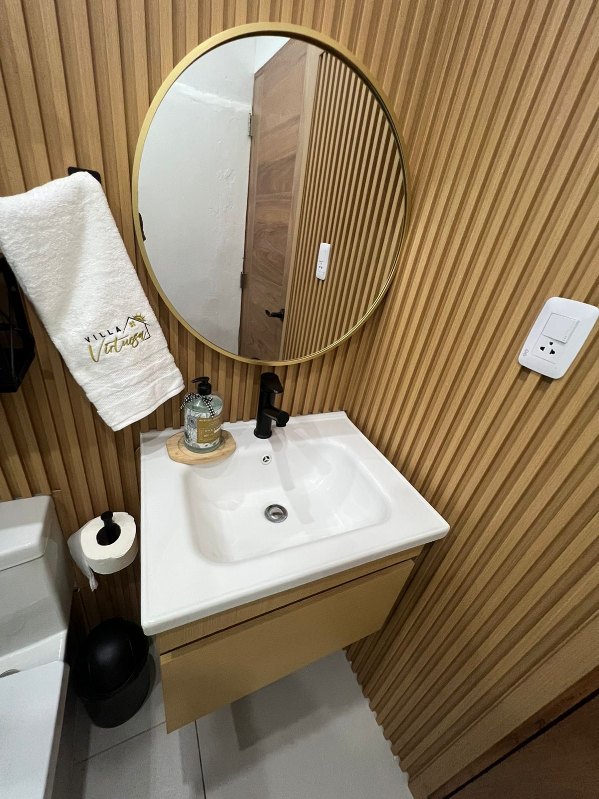 A modern bathroom sink area is presented, featuring a circular mirror framed in gold. A stylish faucet and soap dispenser are arranged on a white countertop, alongside a black trash can. The walls are adorned with vertical wooden paneling, adding texture to the space.