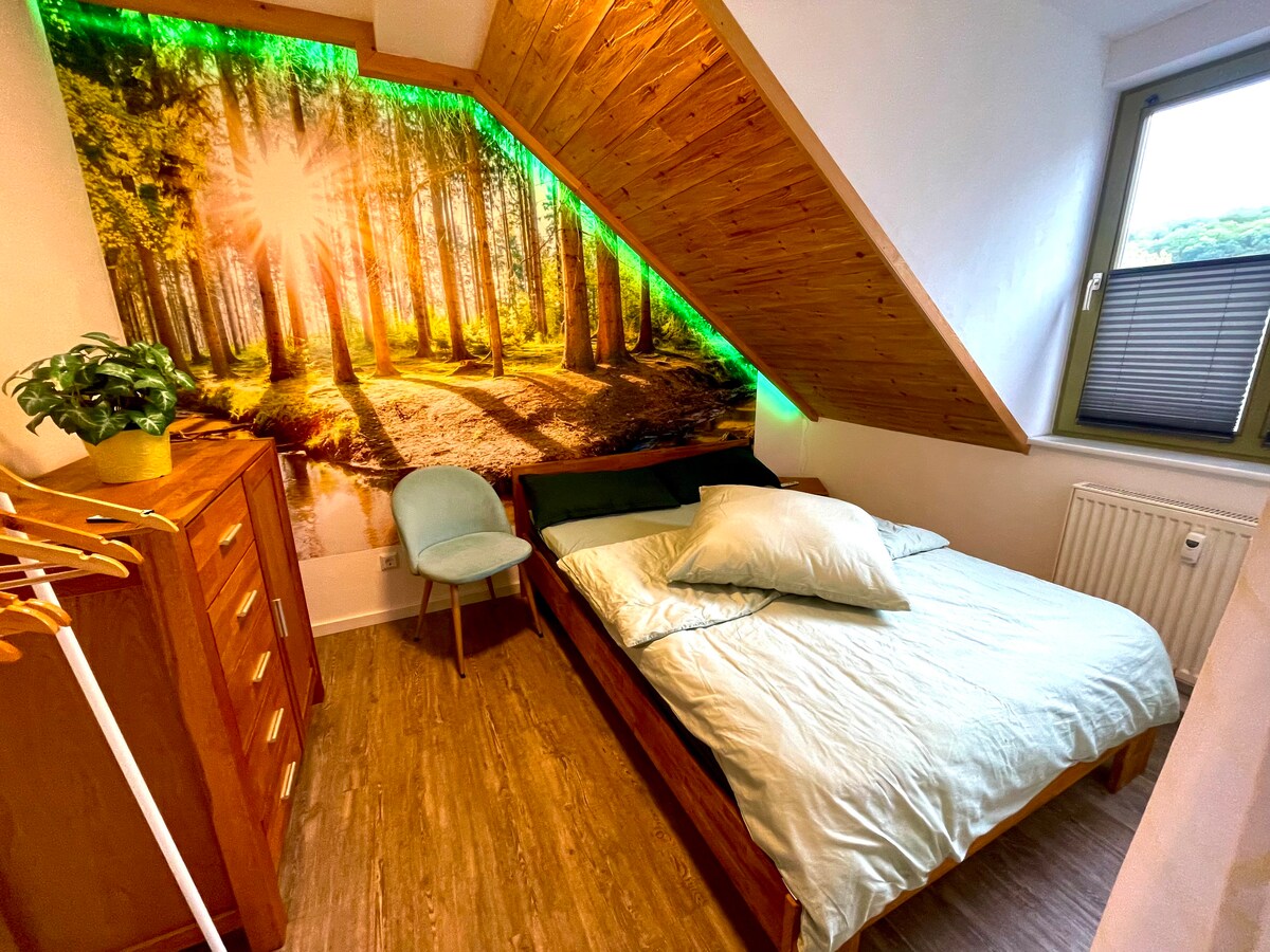 Dormitorio con pared de bosque iluminada. La pendiente está revestida de madera de pino y se adjunta un yeso de yeso alpino a las paredes. Todo lo adecuado para las personas alérgicas y promueve un sueño saludable.
