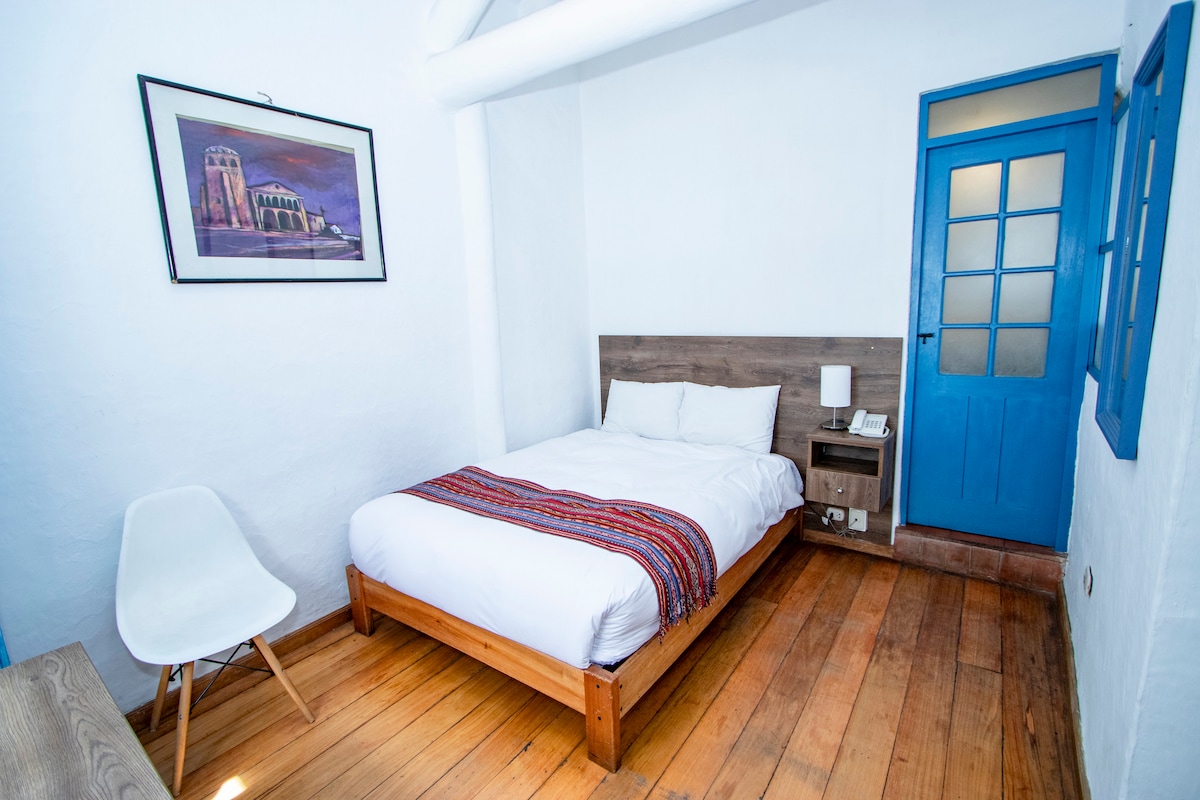 A cozy room features a spacious bed with white linens and a colorful blanket draped across the bottom. Blue doors and trim complement the room's white walls. A minimalist chair adds to the decor, with a framed artwork displayed above the bed.