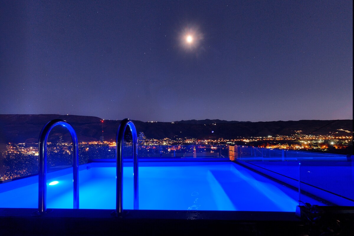 A heated swimming pool is illuminated in soft blue light at night, featuring two silver handrails for easy access. The surrounding area is open, offering views of the distant city lights and mountains, with a clear night sky above.