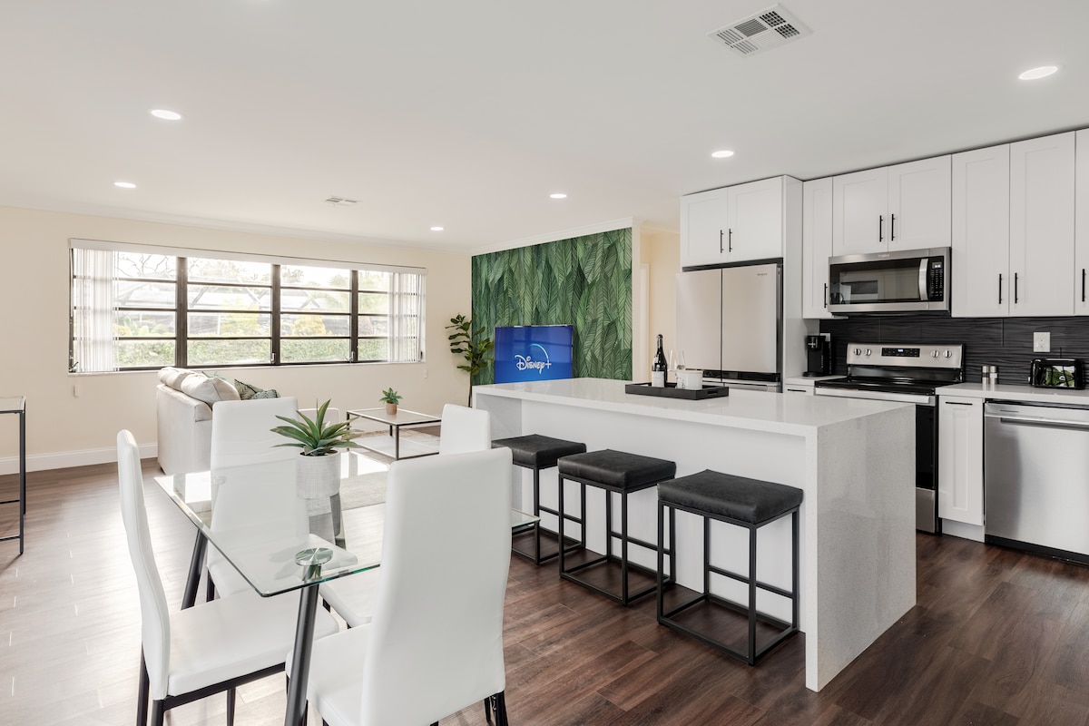 A modern kitchen and dining area are showcased, featuring sleek white cabinetry and stainless steel appliances. A large island provides ample workspace, surrounded by high-backed stools. A dining set is positioned nearby, illuminated by natural light from expansive windows, with leafy green decor enhancing the ambiance.