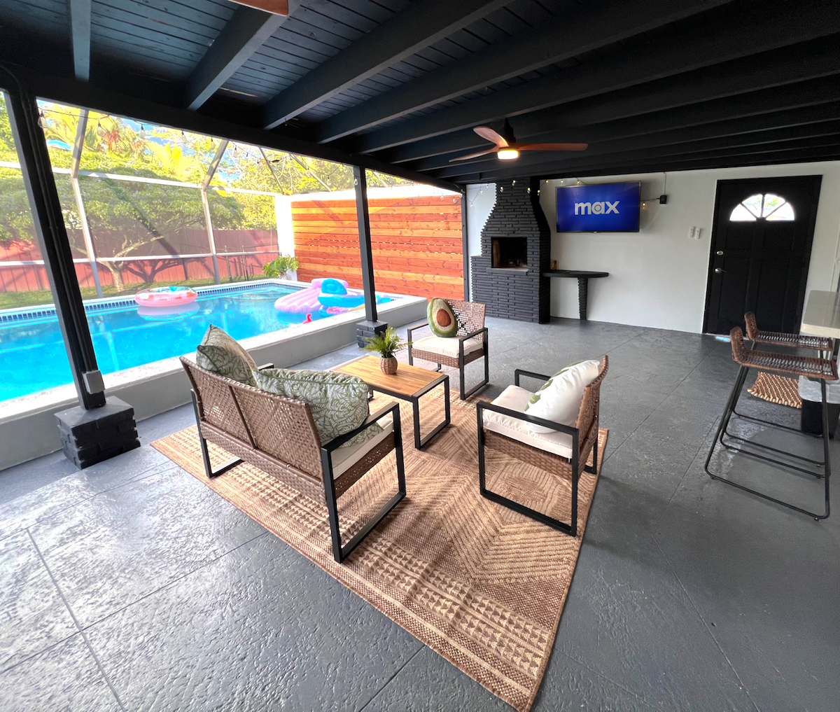 A screened-in outdoor seating area features a cozy arrangement of rattan chairs and a coffee table, positioned on a textured floor. A wall-mounted TV is visible, along with a fireplace. The inviting saltwater pool is just outside, surrounded by greenery.