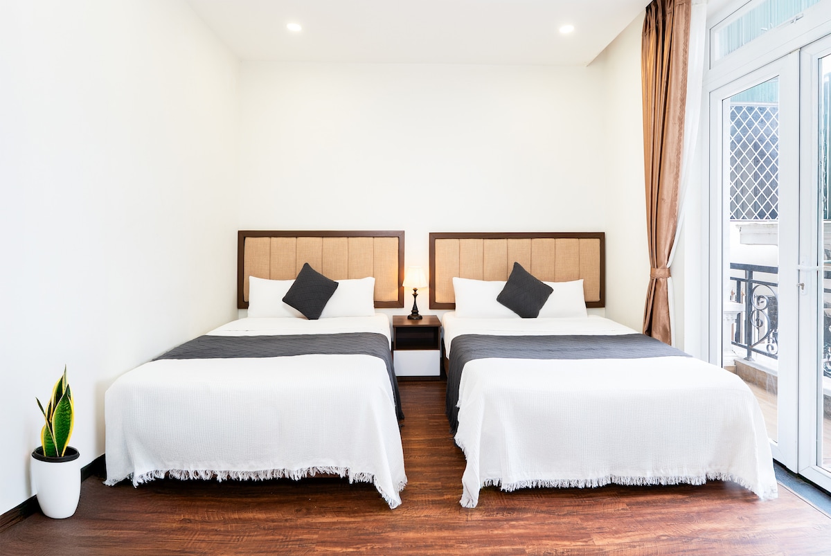 The room is furnished with two spacious beds, each equipped with neutral-colored bedding and decorative pillows. A small bedside table sits between the beds, and a plant is positioned in a white pot. Large windows allow natural light to fill the space.