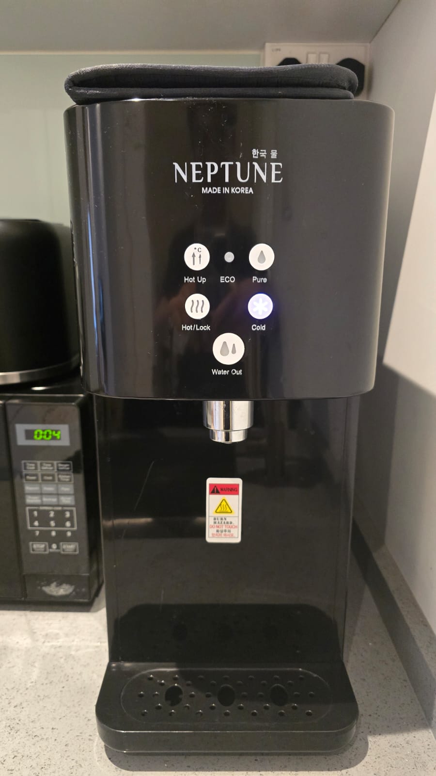 A sleek water dispenser equipped with various settings for heating and cooling, located on the kitchen countertop. Illuminated buttons indicate options for hot, cold, and eco modes. Below the dispenser, a drip tray is visible, designed to catch any spills.