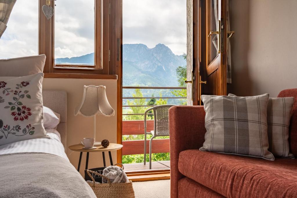 A cozy sleeping area is shown, featuring a spacious bed beside a small nightstand with a lamp. Large windows open to a balcony with mountain views, allowing natural light to fill the room. A sofa and decorative pillows add comfort to the space.